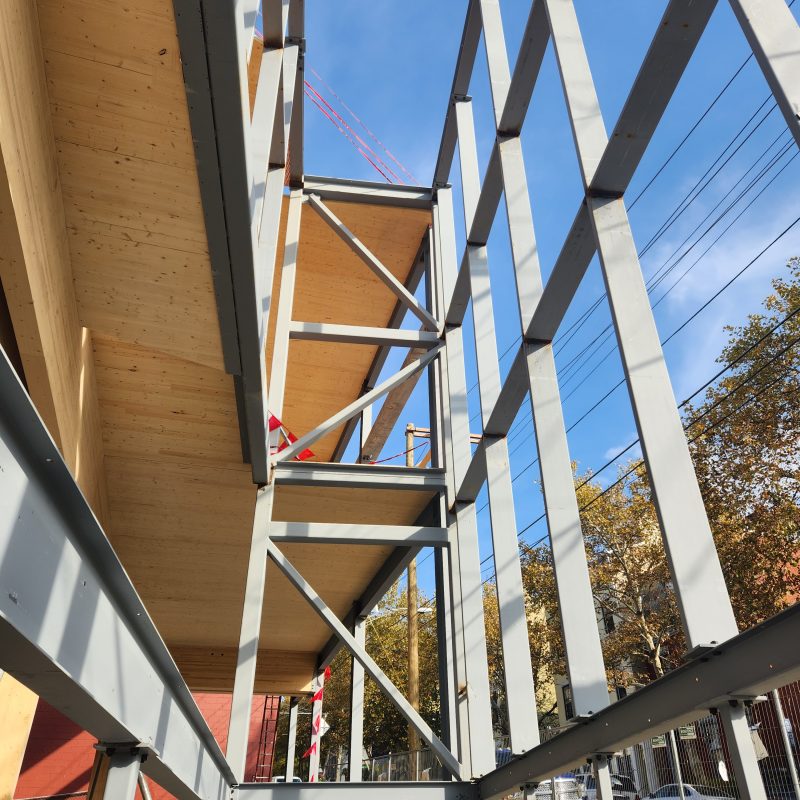 Modern building under construction featuring a steel frame structure with cross-bracing and exposed wooden floors. The construction site is set against a clear blue sky, showcasing urban development with structural engineering techniques.