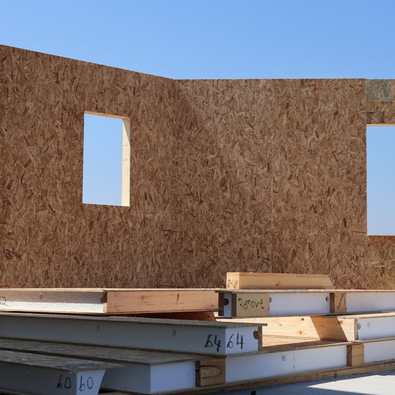 A construction site featuring a partially assembled wall made of Structural Insulated Panels (SIPs). The SIPs are composed of oriented strand board (OSB) with foam insulation sandwiched between the panels, providing structural support and insulation. The panels are labeled and stacked on the ground nearby, ready for installation. Two window openings are visible in the wall, with a clear blue sky in the background.