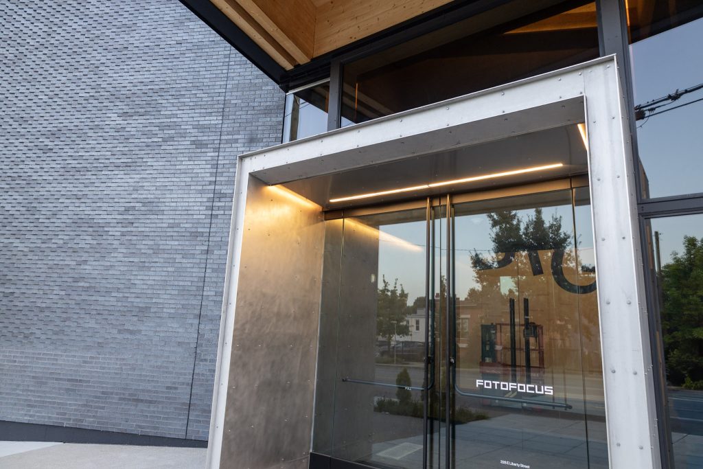 IMG_9930 entrance of the Fotofocus building in Cincinnati, Ohio, with a stainless steel entry portal, glass storefront doors, gray brick facade, and exposed wood ceiling above
